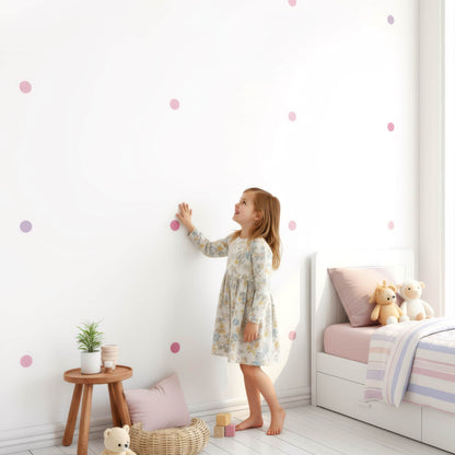 Une petite fille se tient fièrement devant le mur qu'elle a décoré avec des stickers pois rose bonbon, une solution simple pour une décoration de chambre d'enfant personnalisée et créative.