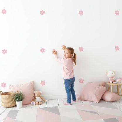 Une petite fille souriante colle des stickers fleurs marguerites roses sur le mur, montrant à quel point cette décoration murale est facile et amusante pour la chambre d'enfant.