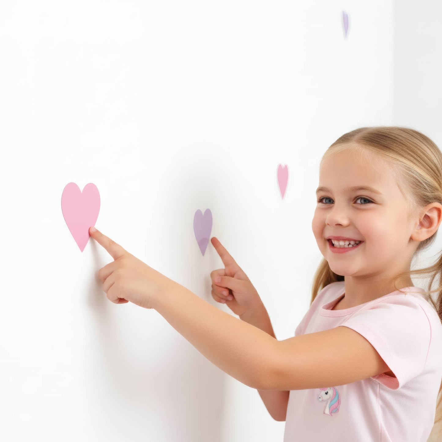 Une petite fille sourit en posant un sticker cœur rose bonbon, montrant à quel point cette décoration murale est amusante et facile à installer dans une chambre d'enfant au Maroc.