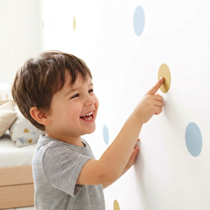 Un jeune enfant souriant pointe du doigt un sticker pois bleu doré sur un mur, mettant en avant la facilité d'utilisation de cette décoration murale pour chambre enfant et bébé.